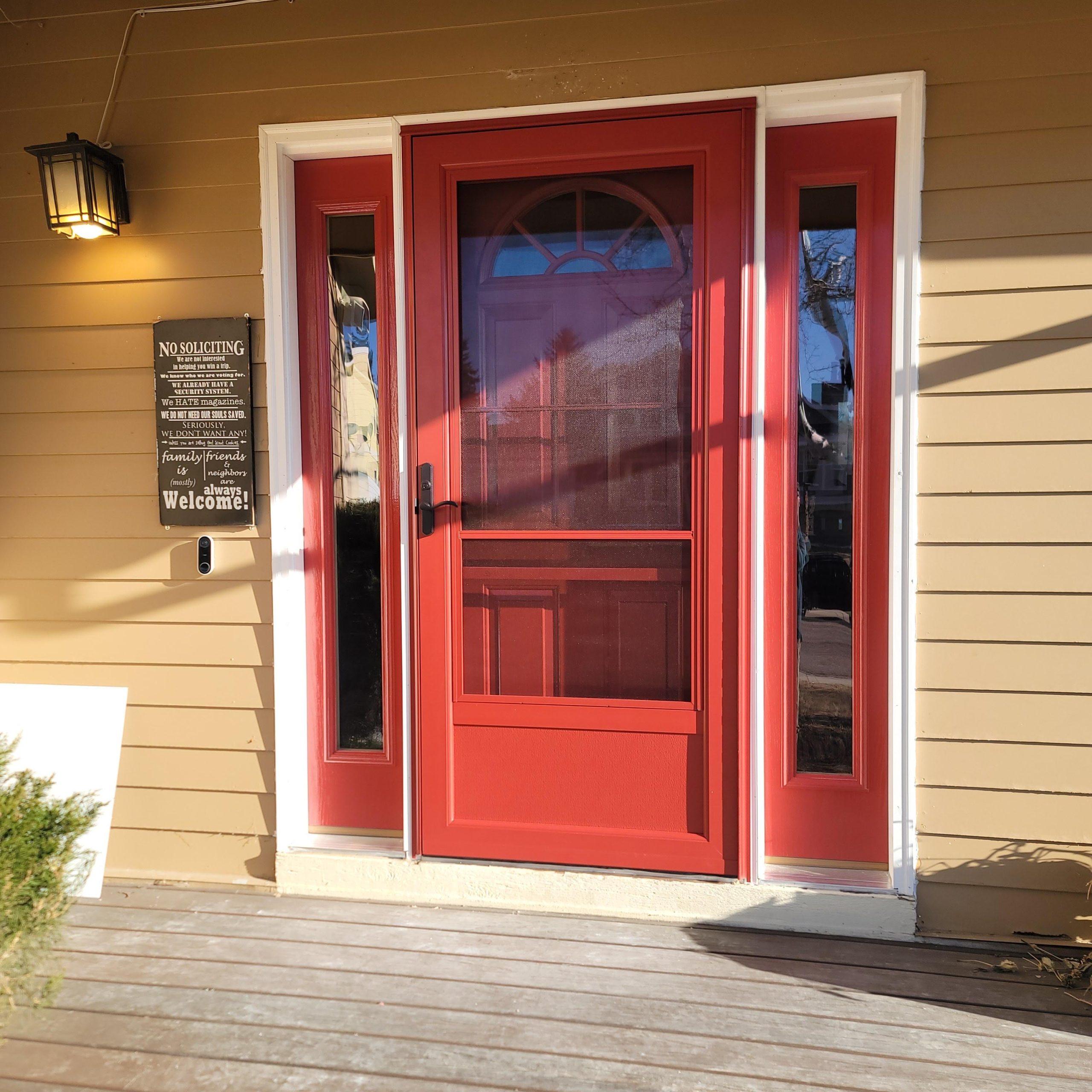 A bright red front door with two vertical glass panels on each side, set in a tan house with white trim. A lantern light and a No Soliciting sign are mounted on the left side of the door. Sunlight casts shadows on the porch.