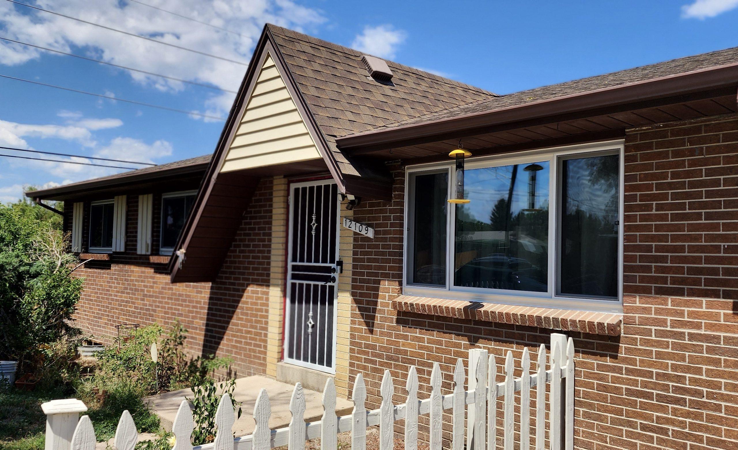 A one-story brick house with a brown roof, white-framed windows, a metal screen door, and a white picket fence in front. The yard has some green shrubs and the sky is clear and blue.