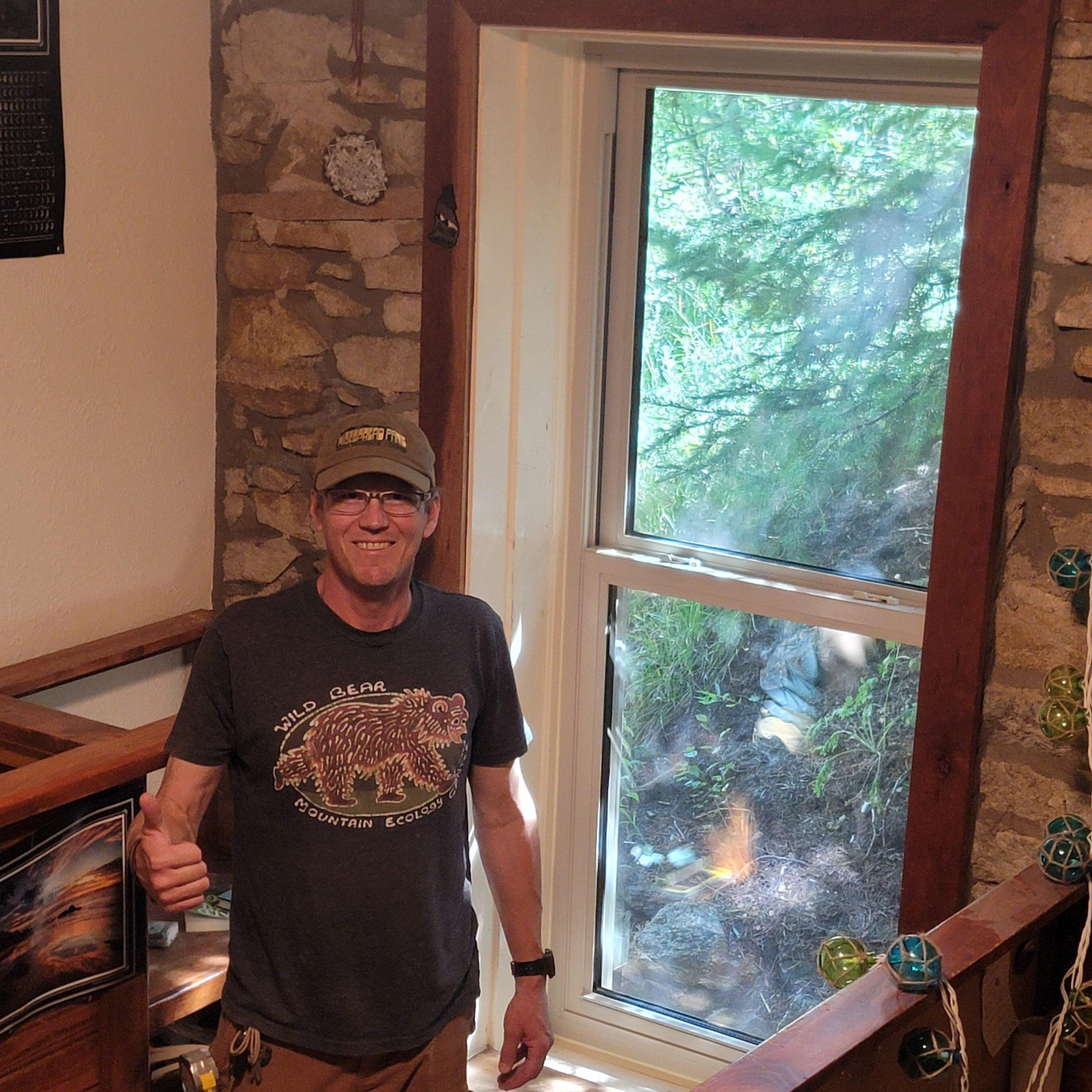 A man wearing glasses, a cap, and a Wild Gear t-shirt stands indoors by a large window with a view of green trees outside, giving a thumbs-up and smiling. The walls are stone, and there are colorful decorations nearby.