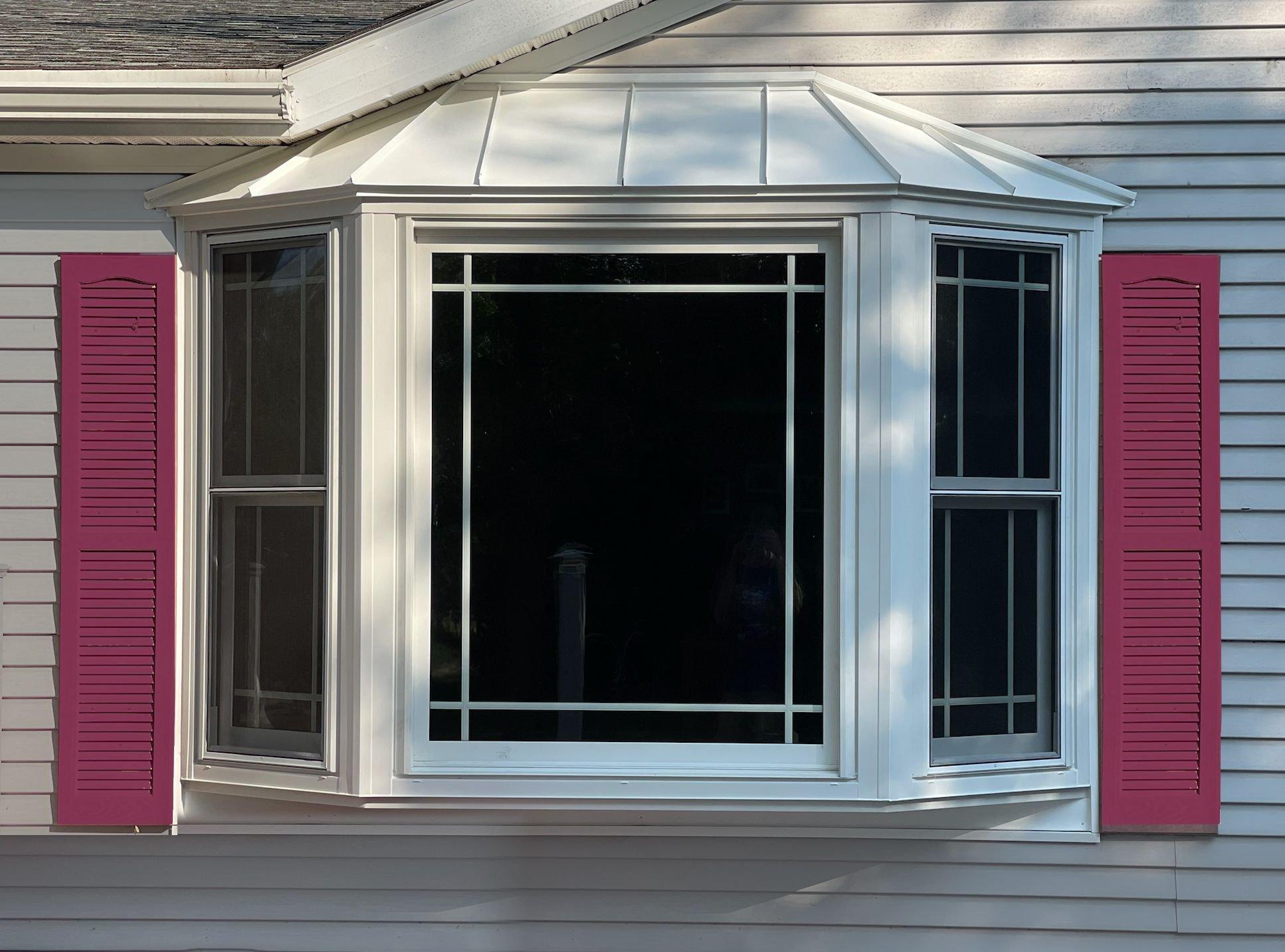 A white bay window with three sections and a metal roof, framed by bright pink shutters, set in the light-colored exterior wall of a house.