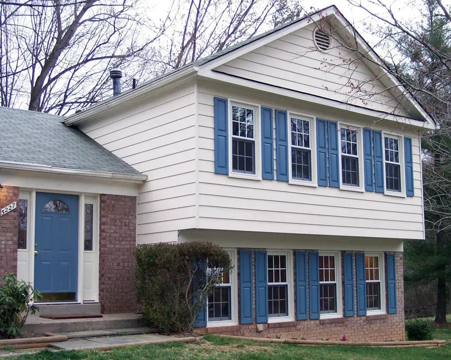 A two-story suburban house with cream siding, blue shutters, and a blue front door. The lower level features brickwork, and there are several windows on both floors. Trees without leaves surround the house.