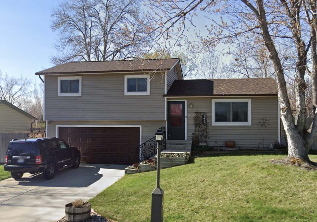 A split-level beige house with a brown garage door, driveway, and SUV parked in front. Leafless trees and a small yard with a lamppost are visible on a clear day.