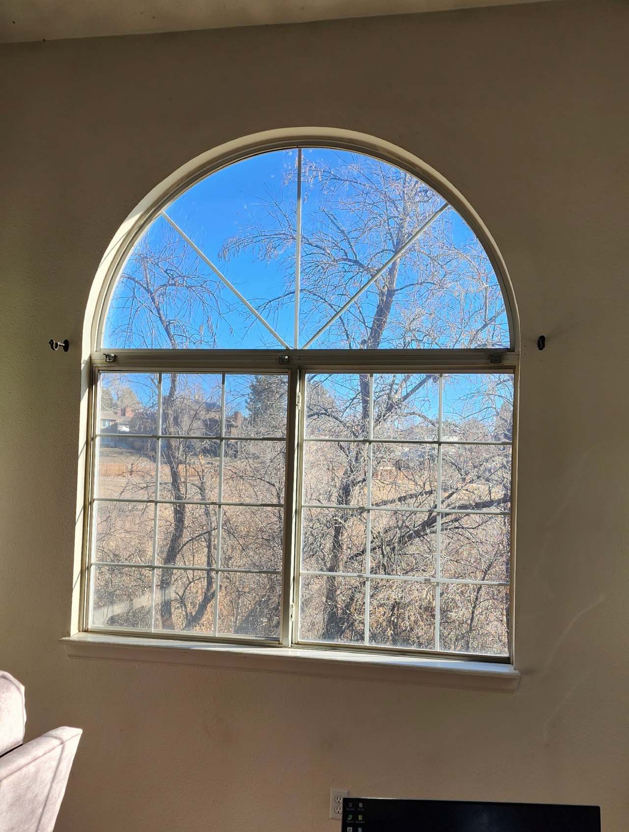A large arched window lets in bright sunlight, revealing a clear blue sky and leafless trees outside. Part of a light-colored wall and a piece of furniture are visible inside the room.